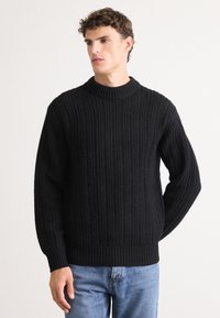 Black knitted sweater with a high neck, ribbed texture, and long sleeves, paired with blue denim jeans. Simple, casual design.