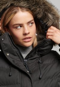 Czarna parka z kapturem wykończonym futrem, zapinana na zamek z przodu i z regulowanymi trokami. Noszona na szarym golfie, z metalowymi akcentami.