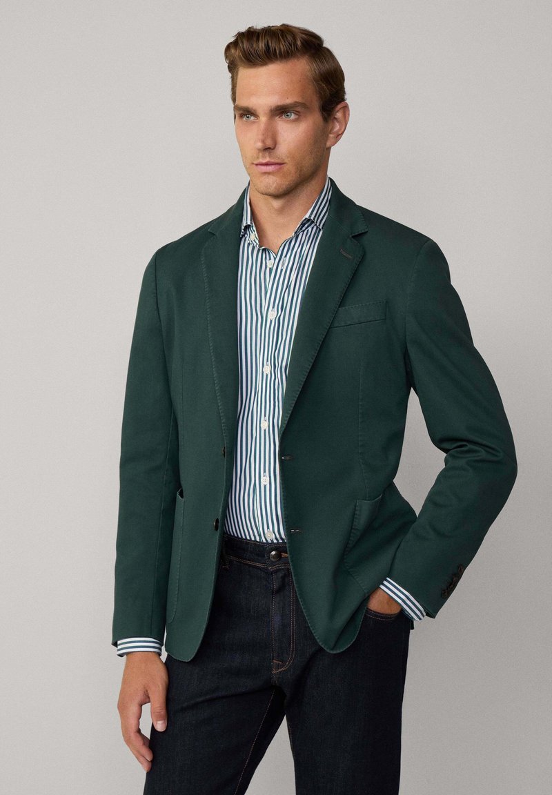 Blazer vert foncé avec une finition texturée, design à un bouton, et deux poches avant porté sur une chemise à rayures bleu et blanche.