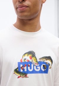 Κοντινό πλάνο λευκού πουκαμίσου με τρία πολύχρωμα ψάρια τυπωμένα γύρω από ένα μπλε τετράγωνο που περιέχει τη λέξη "HUGO" με λευκά γράμματα.