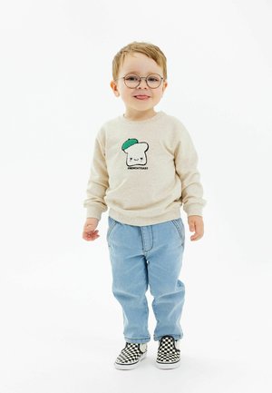 Pulover bej cu un design de pâine desenat animat accentuat în verde, pantaloni din denim albastru deschis și pantofi slip-on în carouri alb-negru.