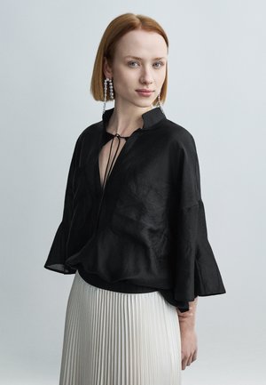 Fekete, könnyű blúz enyhén átlátszó textúrával, harang alakú ujjal és V-nyakkivágással, melyen húzószíjas megkötő található, fehér pliszírozott szoknyával párosítva.
