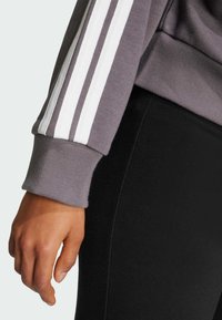 Hihaton harmaa huppari, jossa valkoiset raidat, pitkät hihansuut, yhdistetty mustiin legginsseihin. Sileä materiaali, muotoon ommeltu malli, rento ulkonäkö.