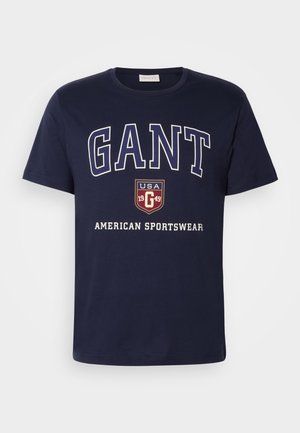 Pils zils kokvilnas T-krekls ar lieliem baltajiem burtiem "GANT" un grafisku logo, kurā norādīts "USA 1949" un "AMERICAN SPORTSWEAR".