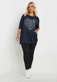 Marinblå oversized topp med korta ärmar som har en stor paljettformad hjärt-design, kombinerad med svarta tajta byxor och vita sneakers.