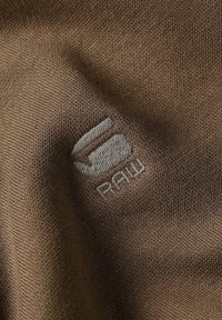 Tessuto marrone con una finitura fine, caratterizzato da un logo "RAW" ricamato di colore grigio chiaro nell'angolo in basso a destra, che mette in risalto elementi di design minimalista.