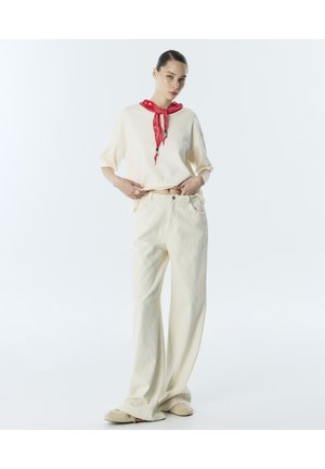 Femme portant un pantalon ample crème, un top oversize assorti, des chaussons beiges et une écharpe rouge nouée autour du cou, debout devant un fond uni.