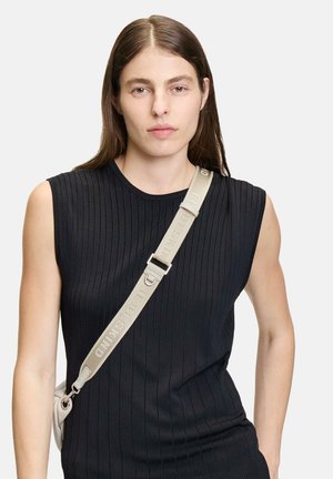 Junge Frau mit glattem braunem Haar trägt ein ärmelloses, schwarzes, geripptes Top und einen beigen, schräg getragenen Taschenriemen mit Text.