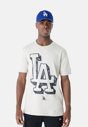 T-shirt in cotone beige con un audace grafico nero "LA". Abbinato a un cappellino blu con lo stesso logo "LA" ricamato sulla parte anteriore.