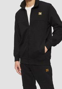 Chaqueta negra con cremallera hecha de tela suave, que presenta un cuello alto, puños acanalados y un pequeño logo dorado en el pecho.