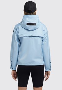Veste imperméable bleu clair avec capuche, dotée d'un patch logo noir et de poignets à bouton-pression. Texture lisse avec un design décontracté.