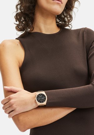 T-shirt marron à manches longues côtelé associé à une montre ronde avec un cadran noir, des accents en or rose et un bracelet noir.