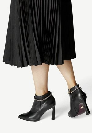 Botines de cuero negro con puntas afiladas, tacones de aguja y acentos decorativos de cadena en el tobillo. Falda negra con pliegues en el fondo.