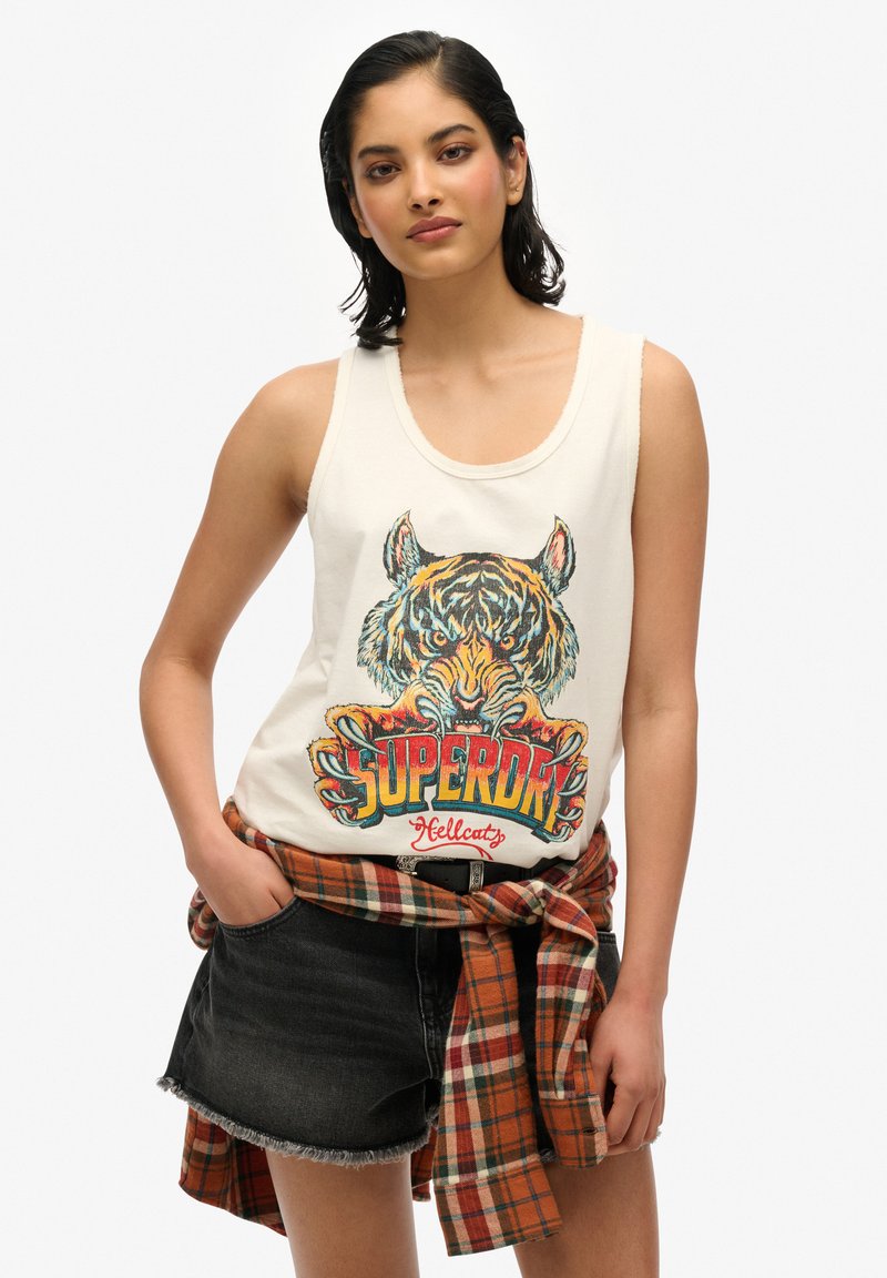 Débardeur blanc avec un motif de tigre coloré et le texte "Superdry Hellcats". Assorti avec un short en denim noir et une chemise à carreaux marron nouée autour de la taille.