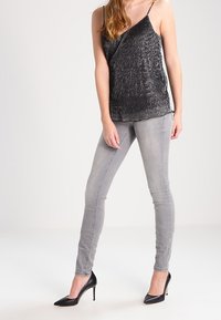 Haut noir pailleté sans manches avec un design croisé, associé à un jean gris clair ajusté et des talons noirs à bout pointu. Textures lisses, style moderne.