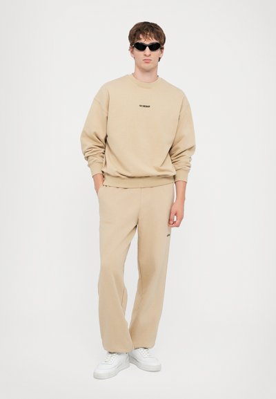 Sweatshirt beige et pantalon de survêtement assorti en tissu doux. Manches longues, poignets côtelés, et baskets blanches pour compléter la tenue. Détail du logo noir.