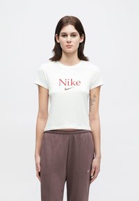 Jovem mulher com cabelo escuro, vestindo uma t-shirt branca cropped da Nike e calças de moletom cor malva, em pé contra um fundo claro e liso.