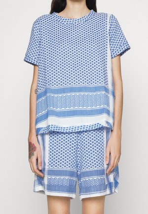 Kobieta ubrana w luźny komplet z krótkim rękawem i szortami z niebiesko-białym geometrycznym wzorem, stojąca na jednolitym tle.