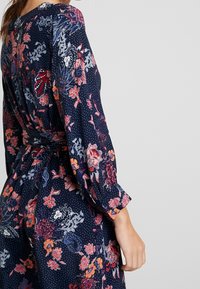 Marinblommig klänning med färgglada mönster i rosa, rött och vitt, med långa ärmar och midja som är åtdragen. Synlig prickig struktur.