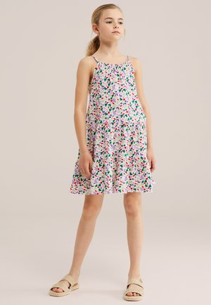 Młoda dziewczyna z blond włosami związanymi w kucyk, ubrana w sukienkę na ramiączkach z warstwowym kwiatowym wzorem i jasne sandały, stoi pewnie.