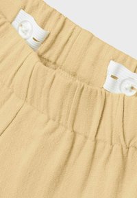 Jasno beżowy elastyczny pasek w talii z marszczoną fakturą i białym zapięciem na guzik. Materiał wydaje się miękki i lekko teksturowany.