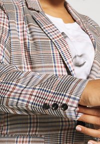 Zbliżenie osoby, która poprawia rękaw wielokolorowej marynarki w kratę z trzema czarnymi guzikami, nosząc białą koszulę z nadrukowanym wzorem twarzy.