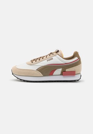 Low-top sneaker med beige ruskind, hvidt stof, brune og røde accenter, brune snørebånd og en sort gummisål.