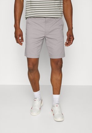 Helles graue Herren-Shorts mit geradem Schnitt, Seitentaschen und umgeschlagenem Saum. Kombiniert mit einem gestreiften grün-weißen Hemd und weißen Sneakers.