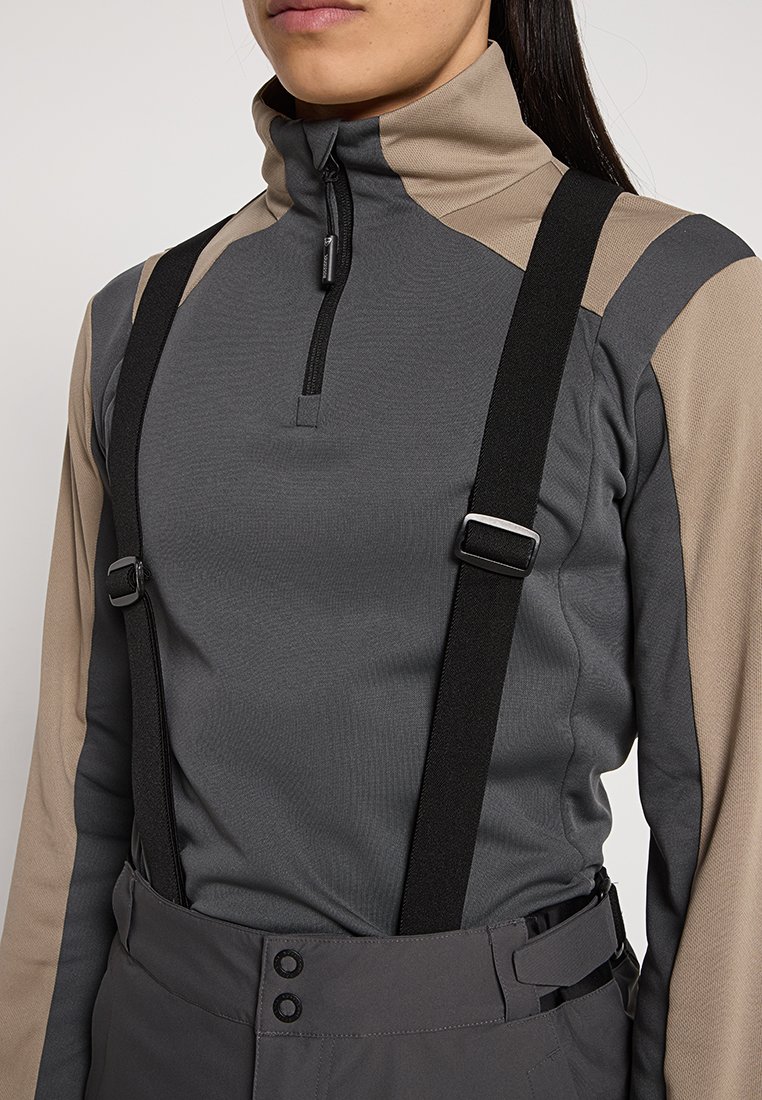 Kobieta ubrana w beżową i szaro-zipowaną bluzę z czarnymi regulowanymi szelkami oraz ciemnymi spodniami, pokazana od ramion do bioder.