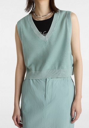 Femme portant un gilet en maille sans manches bleu clair sur une chemise noire, associé à une jupe taille haute bleu clair et des colliers en argent superposés.