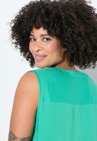 Blusa verde senza maniche con una texture liscia, dotata di scollatura rotonda e di un sottile dettaglio della spalla sul retro, indossata da una persona con capelli ricci.