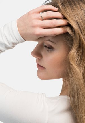 Silberner Zickzackring an einem Finger, Nahaufnahme; das Modell hat lange, wellige braune Haare und trägt ein enges, weißes Langarmoberteil. Einfache Hintergrund.
