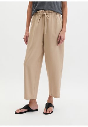 Personne portant un pantalon beige avec un cordon de serrage et des sandales noires à enfiler, debout devant un fond clair uni.