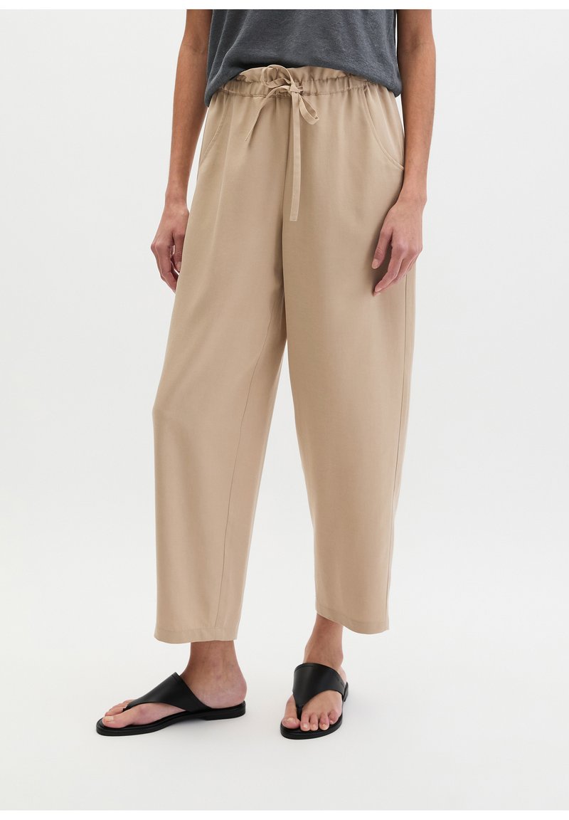 Person wearing beige drawstring pants and black slide sandals, standing against a plain light background.