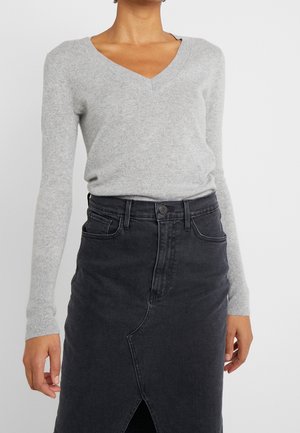 Femme portant un pull gris clair à col en V et une jupe en jean noire taille haute avec une fente à l'avant, debout devant un fond blanc uni.