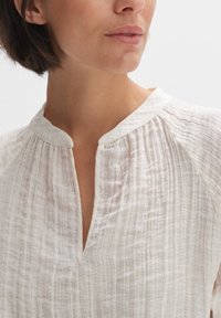 Blusa beige chiaro, con texture e scollatura a chiave, caratterizzata da sottili pieghe verticali e rifiniture a contrasto lungo il colletto e le maniche.