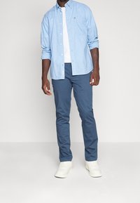 Jasnoniebieska koszula na guziki nałożona na białą koszulkę, zestawiona z niebieskimi spodniami i białymi trampkami. W koszuli widoczny szczegół kieszeni.