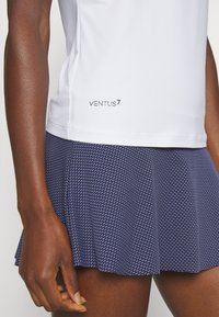 Witte vochtregulerende top met een getextureerde marineblauwe plissérok met een geometrisch patroon en het 'VENTUS7' logo op de zoom.