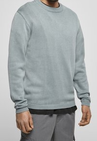 Muž v světle modrém pleteném svetru s kulatým výstřihem přes černou košili a šedých cargo kalhotách, stojící na jednobarevném pozadí.