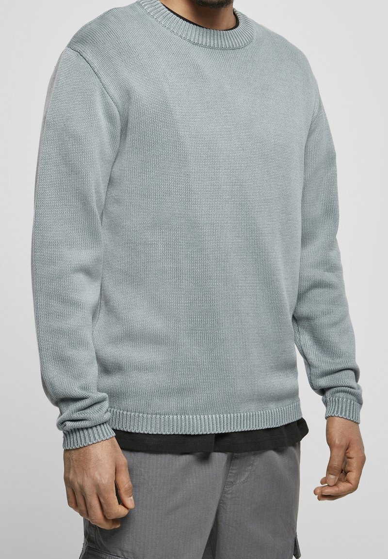 Muž v světle modrém pleteném svetru s kulatým výstřihem přes černou košili a šedých cargo kalhotách, stojící na jednobarevném pozadí.