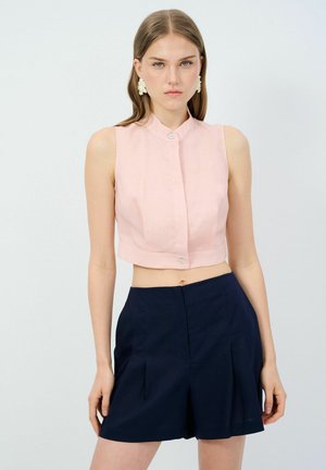Rosa ärmelloses Crop-Top mit hohem Kragen und Frontknopfverschluss, kombiniert mit marineblauen plissierten Shorts. Glatte Stoffstruktur.