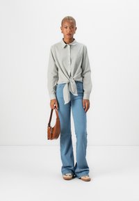 Striped gray button-up shirt tied at the waist, paired with light blue flared jeans and brown crossbody bag, set against a white background.