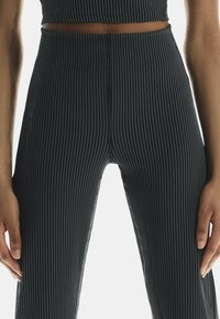 Pantalone s crno-belim prugicama, uskog kroja i glatke teksture, visokog struka, s malim logotipom na strani, prikazani sa rukama na struku.