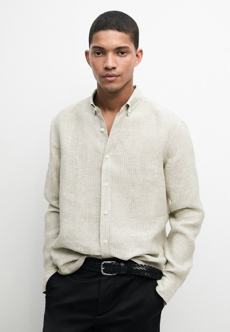 Young man wearing a light beige long-sleeve button-up shirt with black pants and a braided black belt against a plain white background.