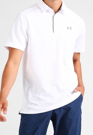 Homme portant un polo blanc à manches courtes avec une fermeture éclair et un logo sur la poitrine, associé à un pantalon bleu.