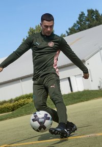 Puma treniņtērps tumši zaļā krāsā ar rozā akcentiem; spēlētājs sit futbolu, tērpies melnās čībās, zāles laukums fonā.