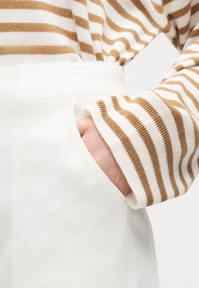 Main dans la poche d'un pantalon blanc, portant un pull à manches longues à rayures horizontales beige et blanc.