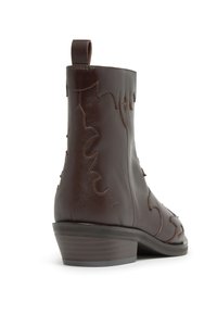 Bota de cuero marrón hasta el tobillo con diseños recortados curvos y costuras en contraste. Cuenta con un tacón bajo apilado y una lengüeta para tirar en la parte trasera.