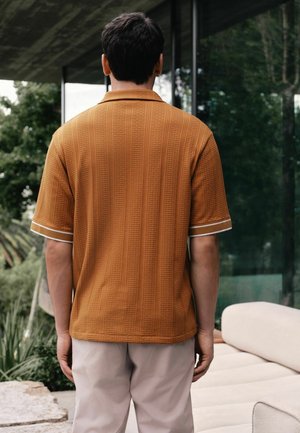 Man wearing a textured, short-sleeve brown shirt and light-colored pants standing indoors beside a beige sofa and glass wall.