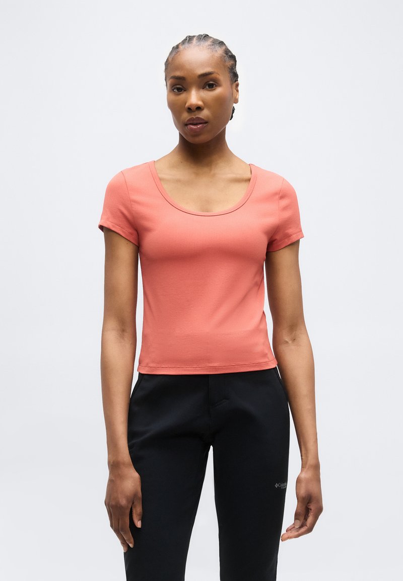 Γυναίκα με πλεξούδες, που φοράει ένα κοραλλί κοντομάνικο μπλουζάκι με στρογγυλή λαιμόκοψη και μαύρο παντελόνι, στέκεται μπροστά σε ένα απλό φωτεινό φόντο.